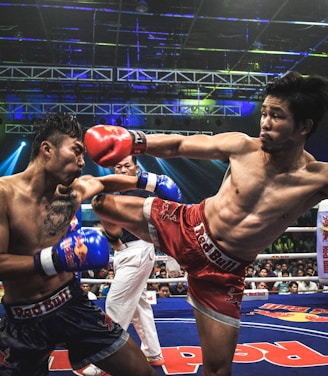 Two athletes are engaged in a dynamic kickboxing match inside a ring, surrounded by a cheering audience. One fighter, wearing red gloves and shorts, executes a high kick towards his opponent, who attempts to block the attack with his arm. The background features colorful lighting and promotional banners.