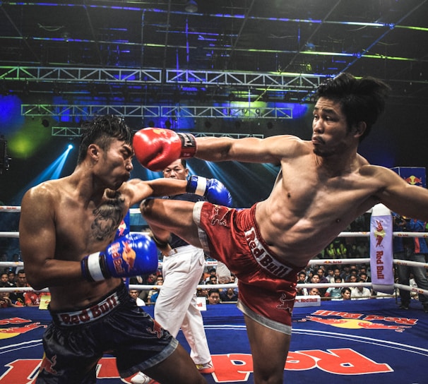 Two athletes are engaged in a dynamic kickboxing match inside a ring, surrounded by a cheering audience. One fighter, wearing red gloves and shorts, executes a high kick towards his opponent, who attempts to block the attack with his arm. The background features colorful lighting and promotional banners.