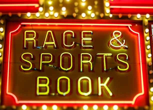 Bright neon lights on a marquee sign display the words 'RACE & SPORTS BOOK'. The sign is surrounded by rows of yellow and white lights against a red background, evoking a vibrant, energetic atmosphere typically associated with casinos or entertainment venues.