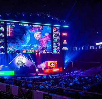 A large esports arena featuring a massive screen displaying a video game, surrounded by sponsor logos and team names. The auditorium is filled with spectators watching the gaming event. Neon blue and purple lights illuminate the stage, creating a vibrant atmosphere.