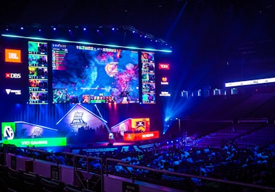 A large esports arena featuring a massive screen displaying a video game, surrounded by sponsor logos and team names. The auditorium is filled with spectators watching the gaming event. Neon blue and purple lights illuminate the stage, creating a vibrant atmosphere.