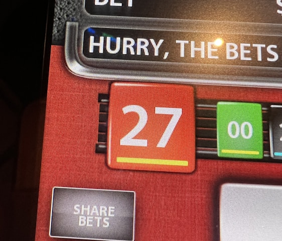 A digital display screen showing a casino betting interface, with text prompting to hurry bets and a highlighted number 27 in red. There are options for betting amounts and a 'share bets' button.