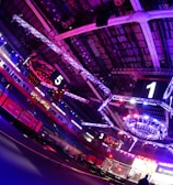 An indoor arena with vibrant purple and blue lighting, featuring large numbered screens suspended from the ceiling. The venue has a high ceiling with intricate metal truss structures and hanging lights, creating a futuristic and energetic atmosphere.