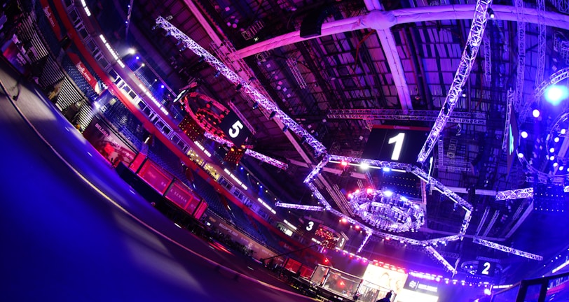 An indoor arena with vibrant purple and blue lighting, featuring large numbered screens suspended from the ceiling. The venue has a high ceiling with intricate metal truss structures and hanging lights, creating a futuristic and energetic atmosphere.