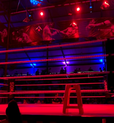 An empty boxing ring is illuminated by dramatic red and blue spotlights inside a dimly-lit arena. A small stool is placed in the center of the ring, surrounded by ropes. The arena is adorned with banners depicting action scenes, and a few silhouettes of spectators can be seen in the background.