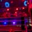 A brightly lit boxing or wrestling ring with vivid red and blue lights. Banners with images of fighters hang from the ceiling. The ropes of the ring are prominently visible, and a stool is placed inside the ring. The arena is filled with an audience, silhouetted against the colorful lighting.