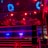 A brightly lit boxing or wrestling ring with vivid red and blue lights. Banners with images of fighters hang from the ceiling. The ropes of the ring are prominently visible, and a stool is placed inside the ring. The arena is filled with an audience, silhouetted against the colorful lighting.
